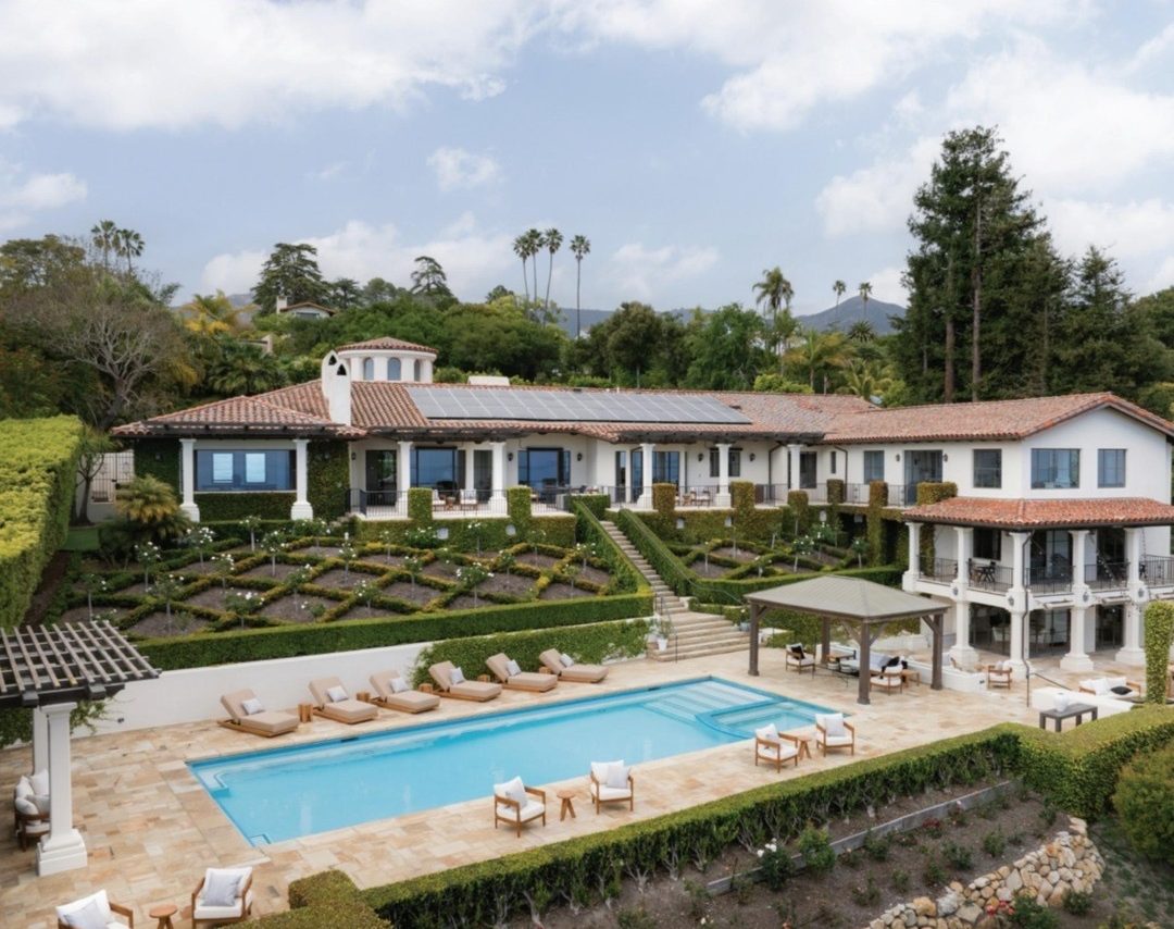 Aerial photo of the estate at 937 Cima Linda Lane: a large white house with a tile roof, terraced gardens and a pool, on a wooded hillside.