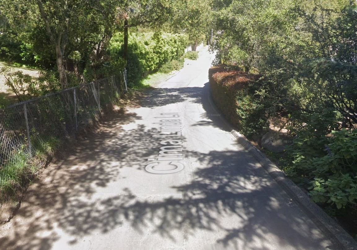 Cima Linda Lane: a narrow paved lane between a chain-link fence and a hedge, under tree cover. Google Street View, March 2019.