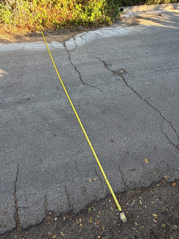 A yellow tape measure stretched across the full width of Cima Linda Lane, from pavement edge to pavement edge.